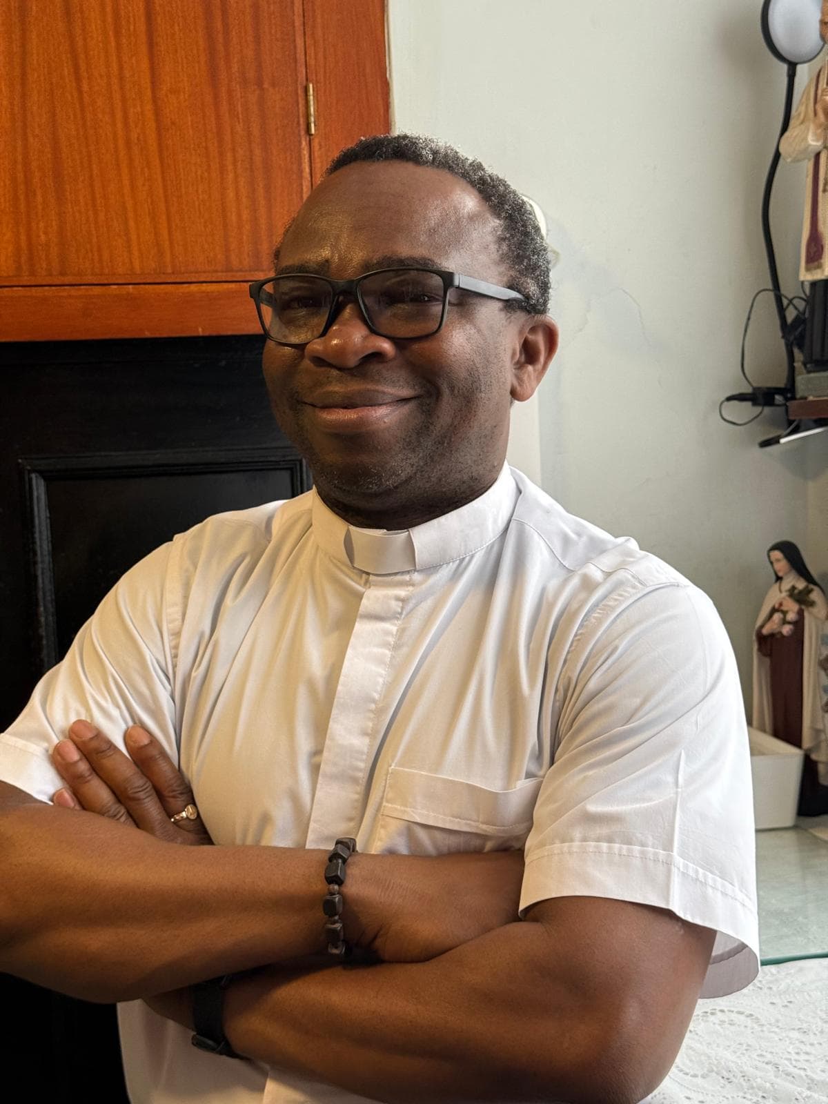 Fr Kenneth Iwunna, Assistant Priest, smiling with arms folded in a white clerical shirt in the parish office